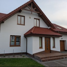 Alpi-Nika Artur Wójtowicz - Biały dom jednorodzinny z brązowym dachem i stolarką. Elewacja po malowaniu, widoczne detale wykończenia i otoczenie domu. Ujęcie z poziomu terenu.