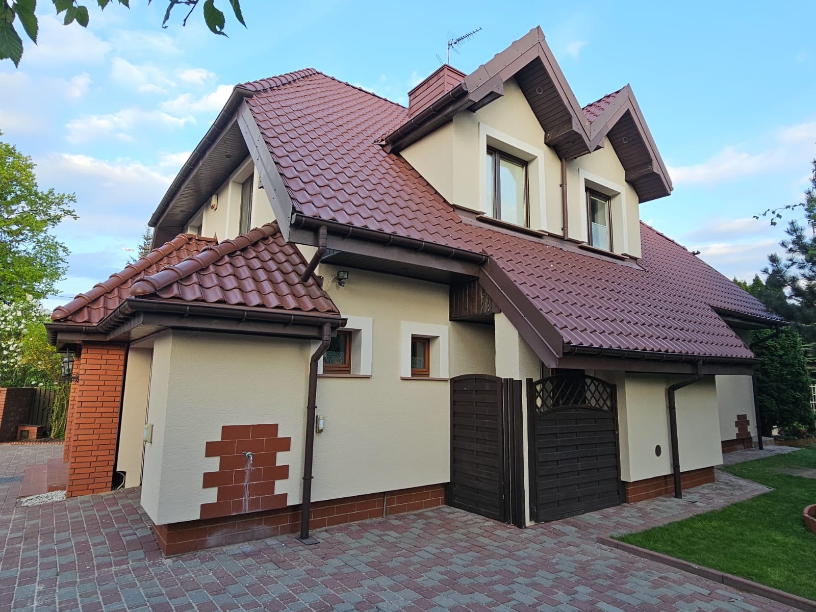 Dwupiętrowy dom z beżową elewacją, brązowym dachem i elementami dekoracyjnymi z cegły, widziany z perspektywy chodnika.