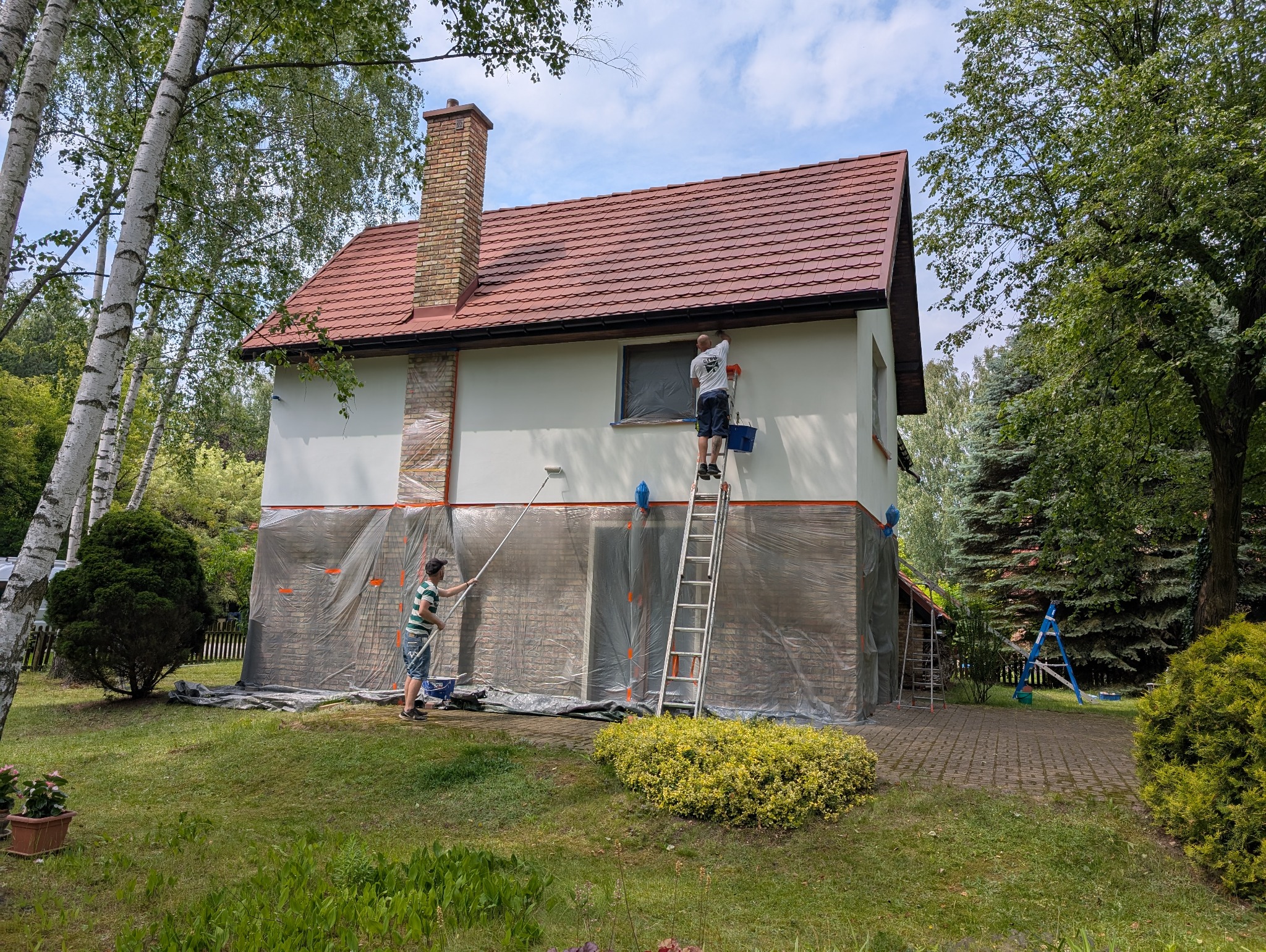 Dwóch mężczyzn maluje elewację domu jednorodzinnego. Jeden stoi na drabinie, drugi używa wałka z ziemi. Część elewacji zabezpieczona folią. Widok z ogrodu.