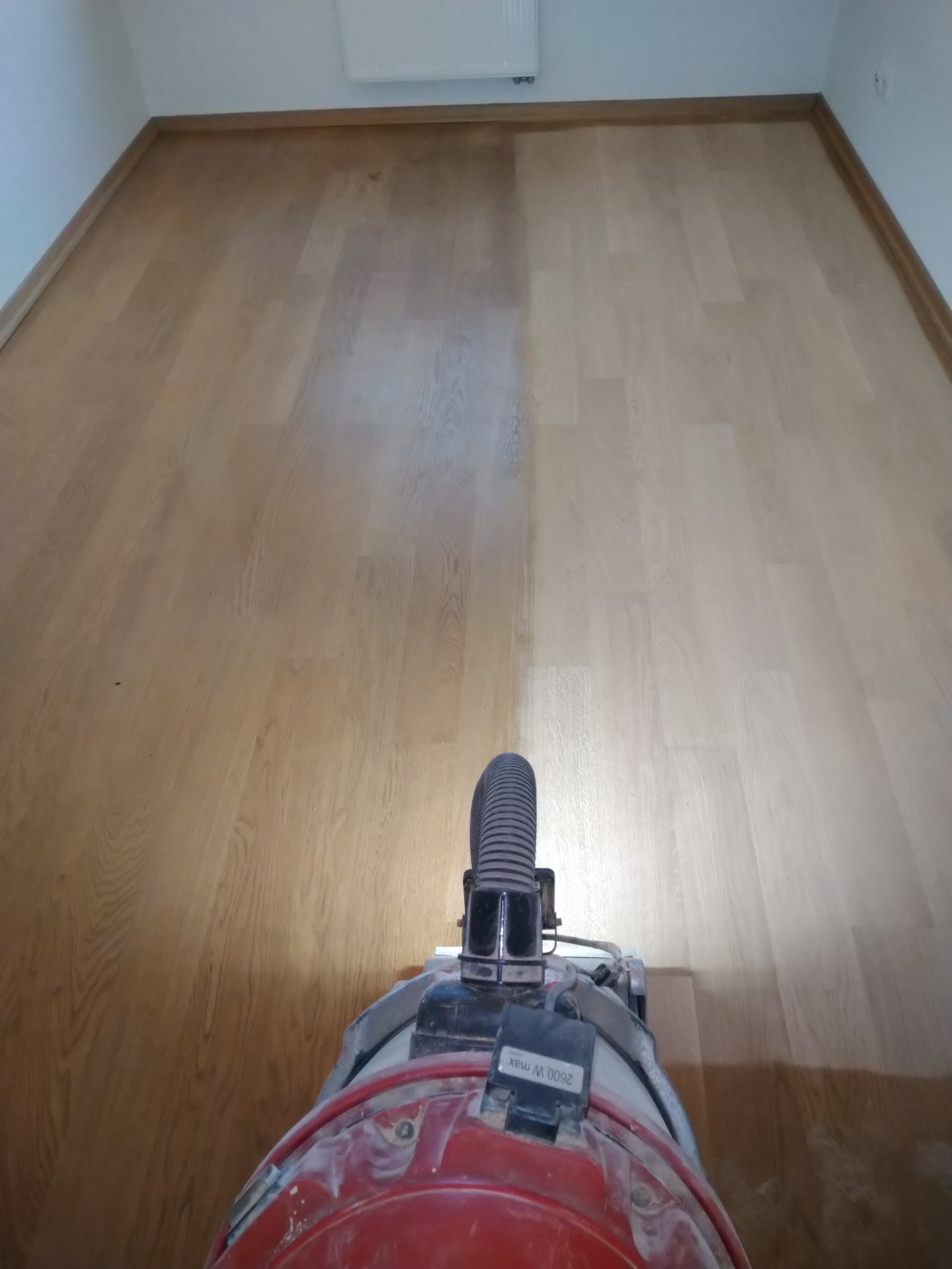 Parkiet po częściowym cyklinowaniu. Widoczna różnica w kolorze i strukturze drewna. Perspektywa z góry na maszynę do cyklinowania i podłogę.