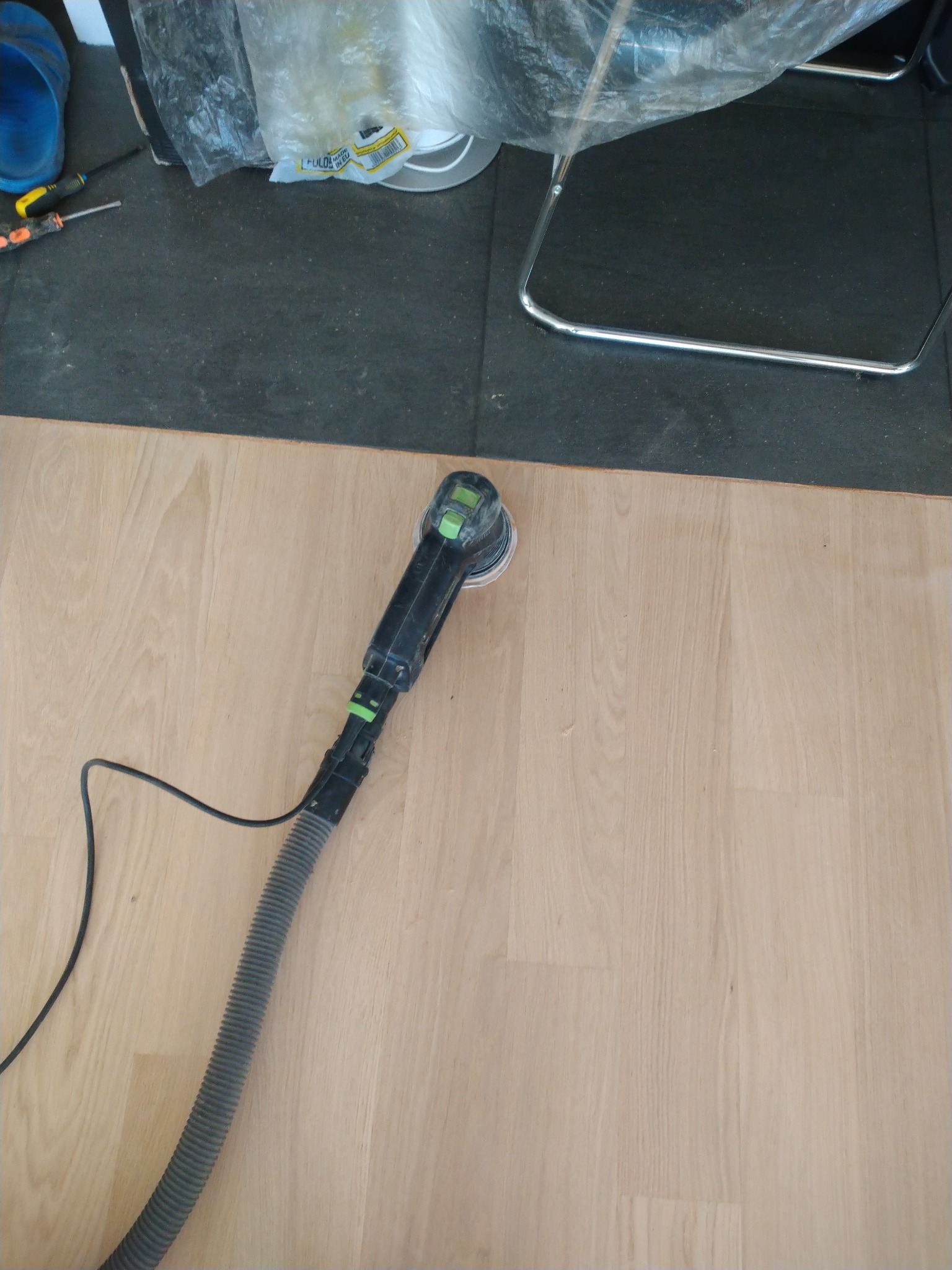 Szlifowanie jasnego parkietu szlifierką oscylacyjną z odkurzaczem. Widoczne narzędzia pracy i fragment krzesła. Ujęcie z góry, bliski plan.