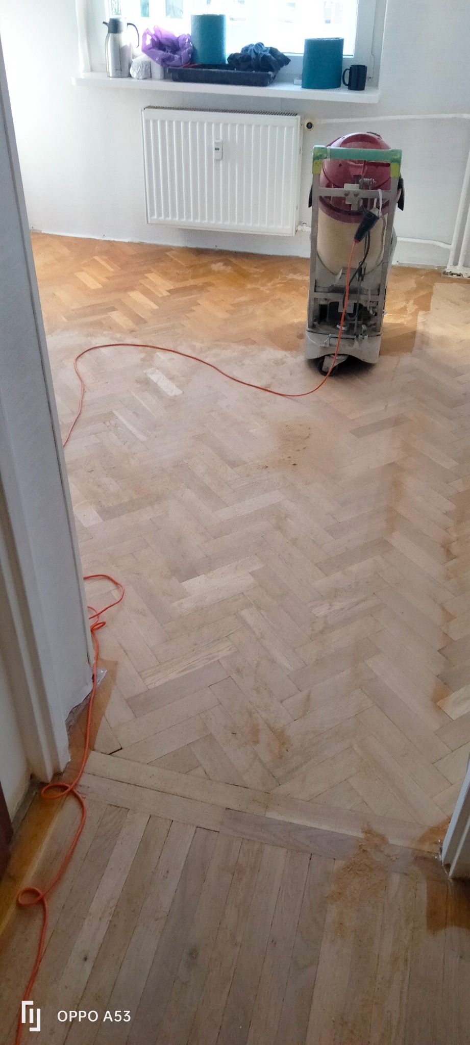 Parkiet w jodełkę częściowo wycyklinowany, widoczna maszyna do cyklinowania. Podłoga w trakcie renowacji, z widocznymi różnicami w odcieniach drewna.