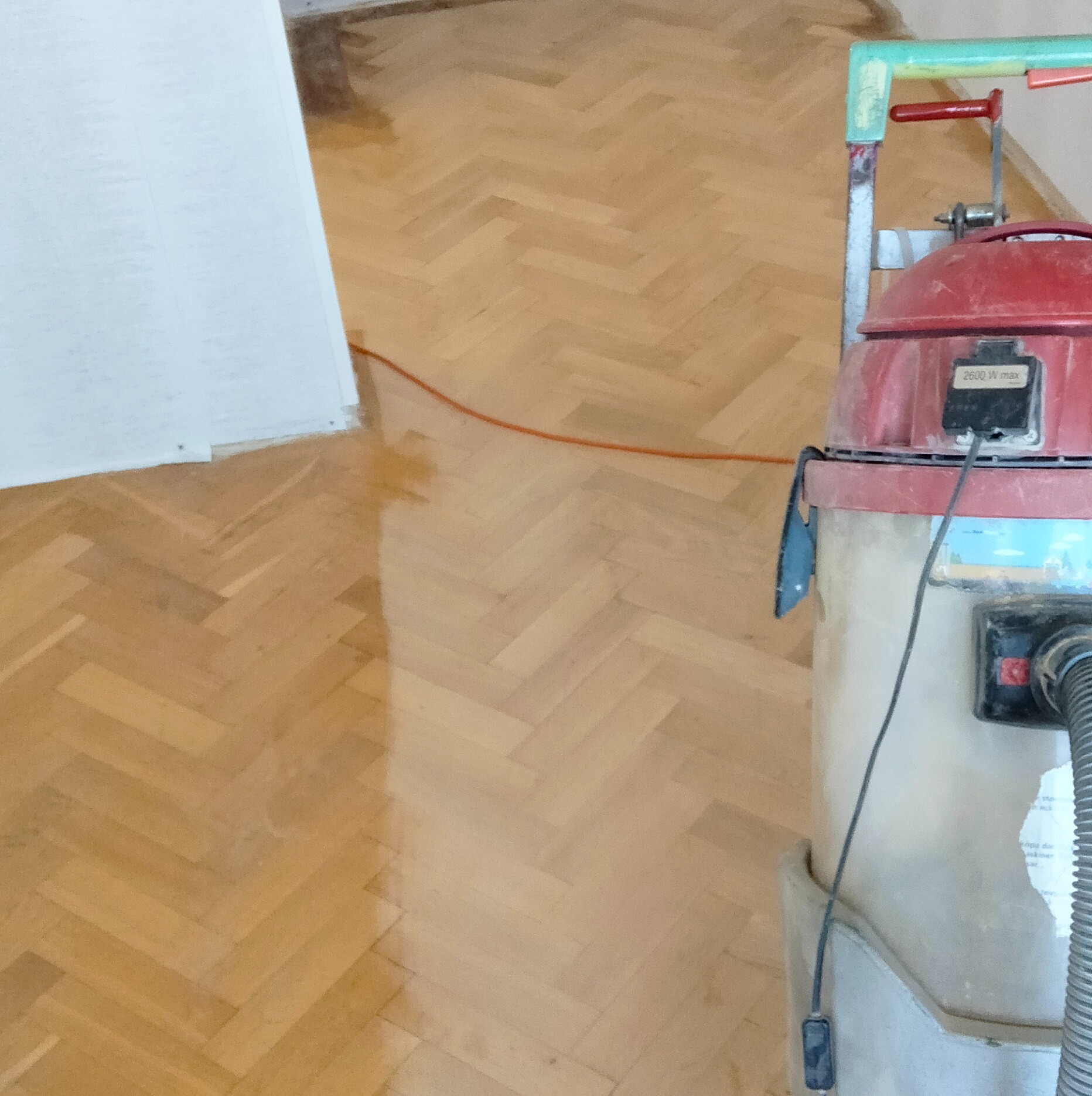 Parkiet w jodełkę po renowacji, częściowo błyszczący, z maszyną do polerowania. Widoczny kabel zasilający i fragment ściany. Jasne, naturalne oświetlenie.