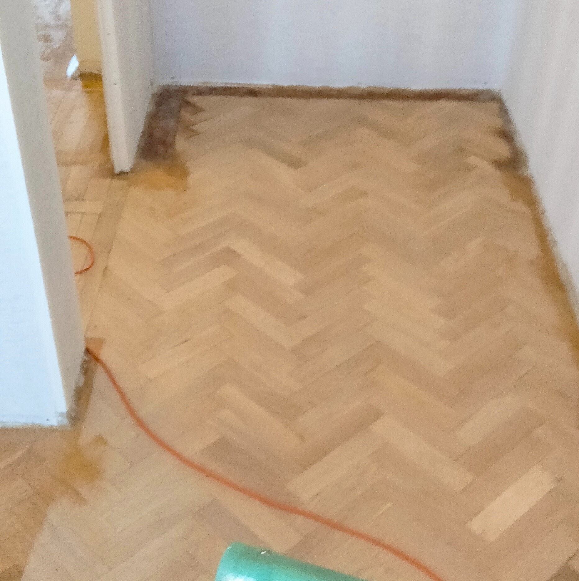 Cyklinowanie parkietu ułożonego w jodełkę. Widoczny fragment cykliniarki i kabel. Częściowo obrobiony parkiet przy ścianach. Podłoga po renowacji.