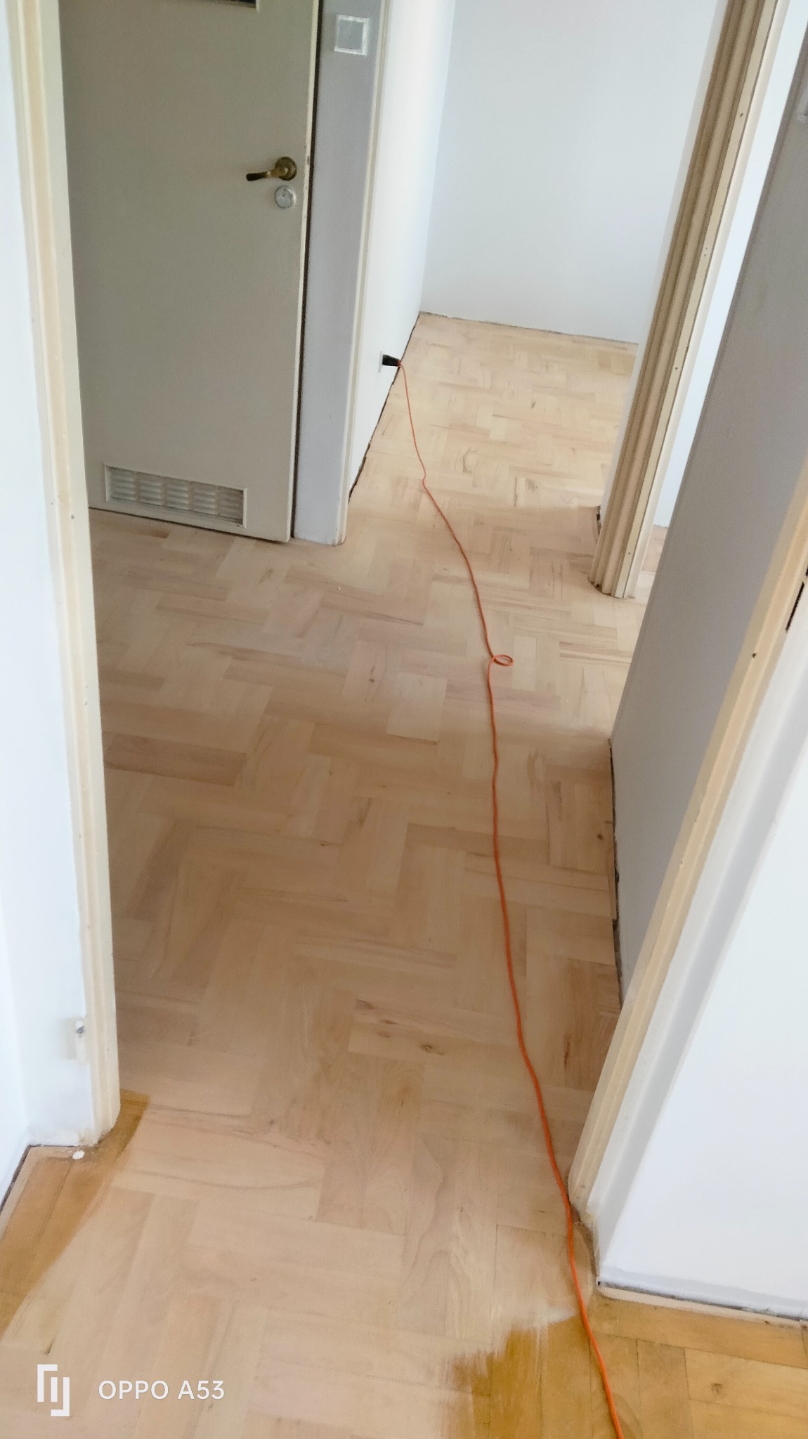 Jasny, odnowiony parkiet ułożony w jodełkę, widoczny z perspektywy wnętrza. Pomarańczowy kabel na podłodze, białe ściany i drzwi. Widok na sąsiednie pomieszczenie.