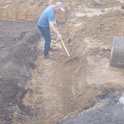 Zielony ogród - Mężczyzna wyrównuje teren grabiami w wykopie, przygotowując podłoże pod ogród. Na drugim planie hałda ziemi i zieleń drzew.