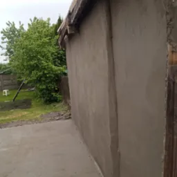 Świeżo otynkowana ściana budynku z widocznym fragmentem dachu i podmurówką, na tle zieleni ogrodowej. Prace wykończeniowe w toku, surowy stan.