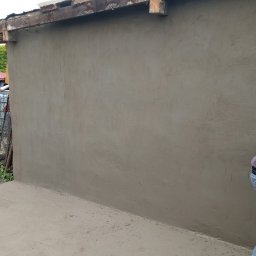 Zielony ogród - Świeżo otynkowana ściana budynku z widocznym fragmentem dachu i pracownikiem zacierającym tynk pacą. Ściana szara, tynk jeszcze wilgotny. W tle zieleń i zabudowania.