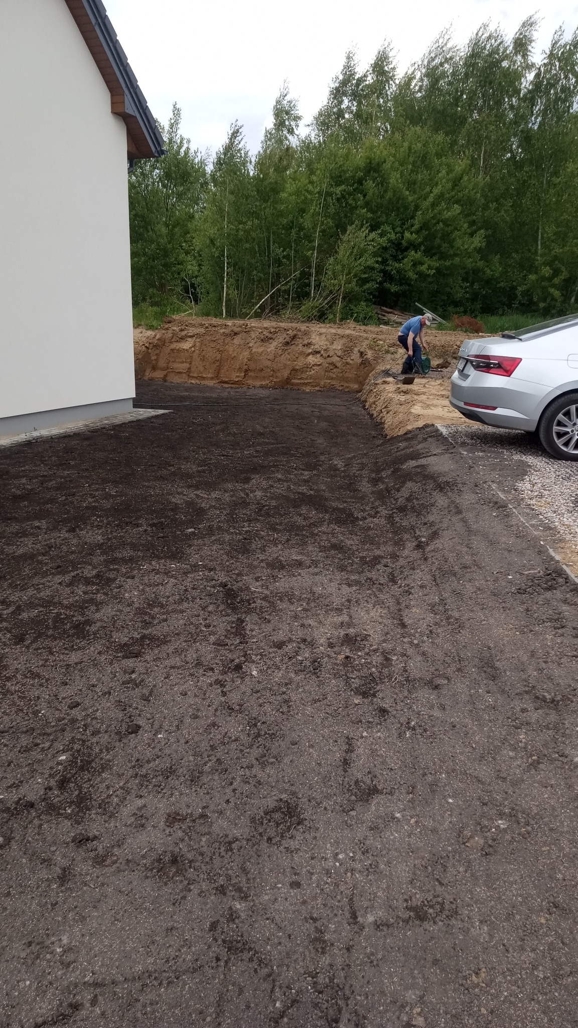 Przygotowany teren pod trawnik z widoczną warstwą ziemi, pracownik wyrównujący teren oraz fragment domu i samochodu w tle, sugerujące świeżo zagospodarowaną przestrzeń.