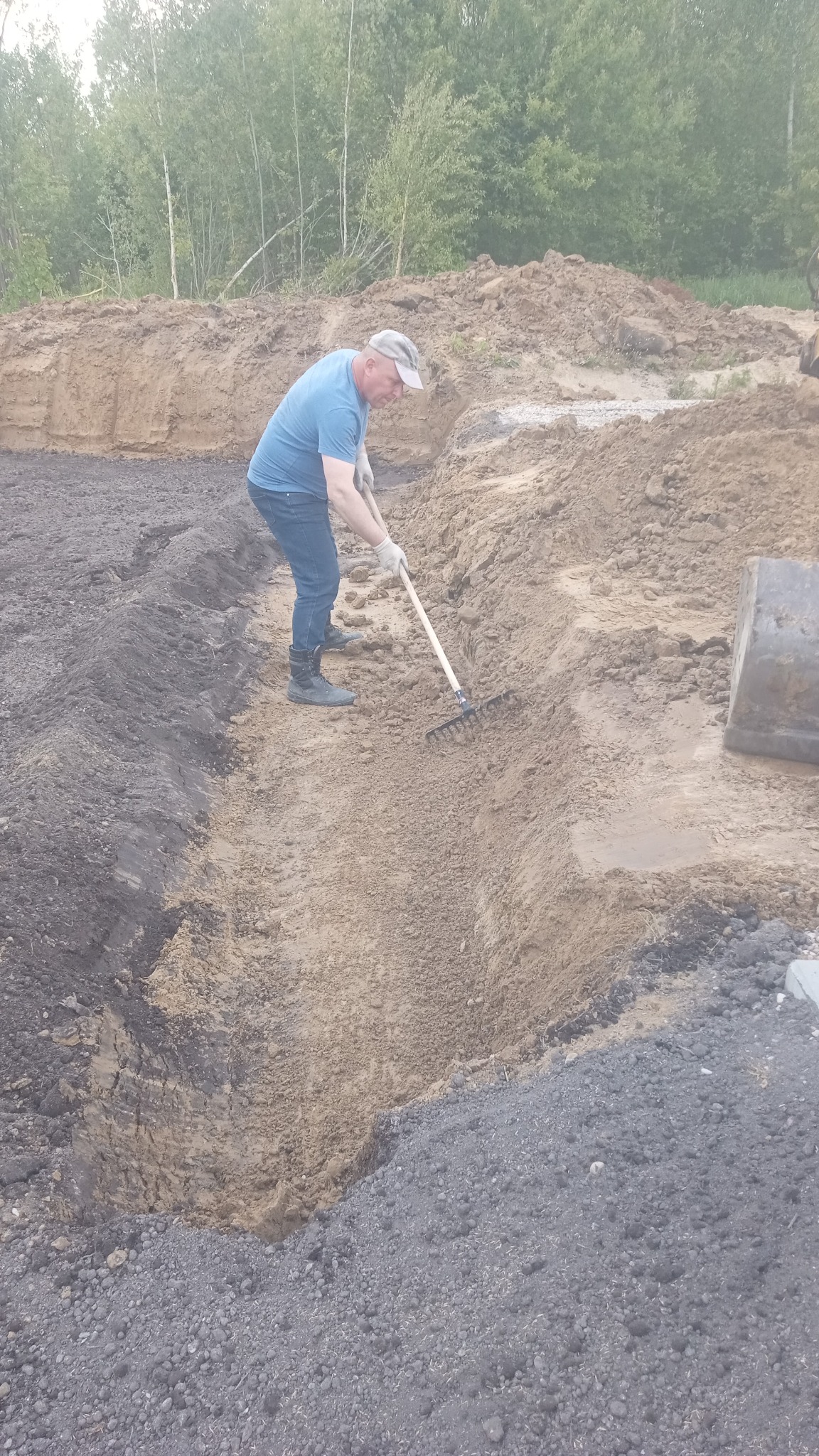 Mężczyzna wyrównuje teren grabiami w wykopie, przygotowując podłoże pod ogród. Na drugim planie hałda ziemi i zieleń drzew.