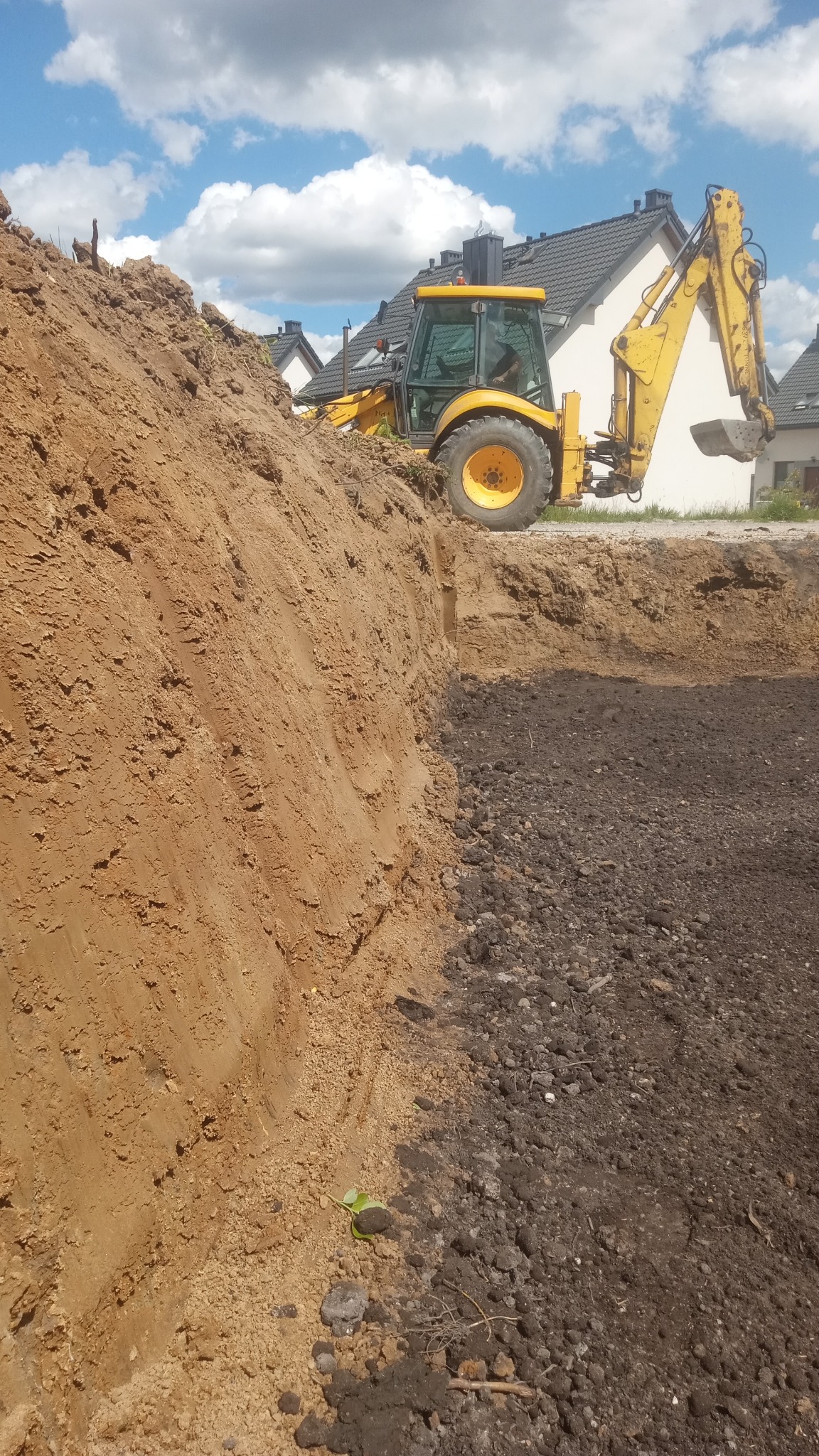 Żółta koparko-ładowarka na skraju wykopu, na tle domów i błękitnego nieba z chmurami. Widoczna ściana wykopu z ziemi i gruzu.