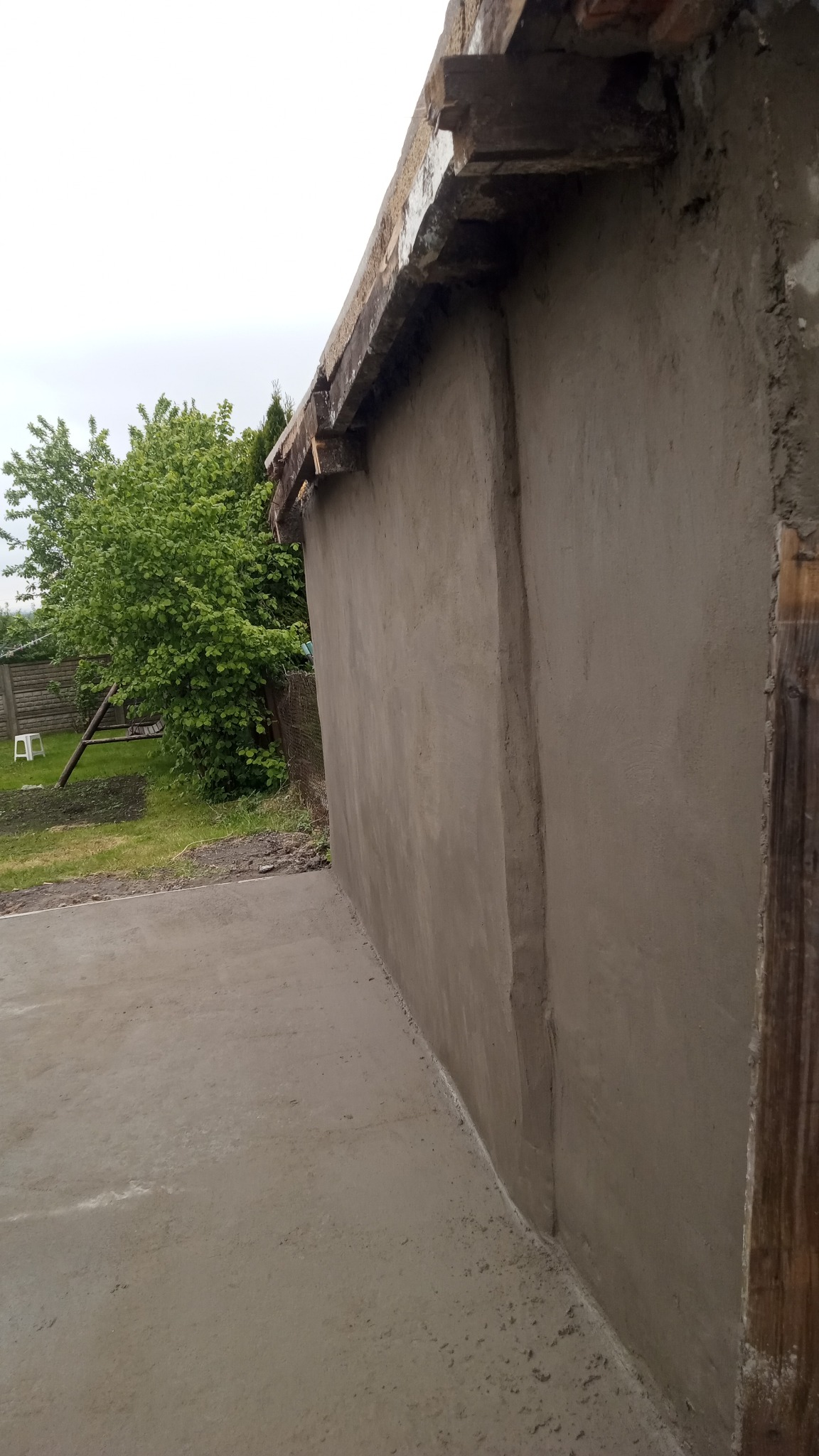 Świeżo otynkowana ściana budynku z widocznym fragmentem dachu i podmurówką, na tle zieleni ogrodowej. Prace wykończeniowe w toku, surowy stan.