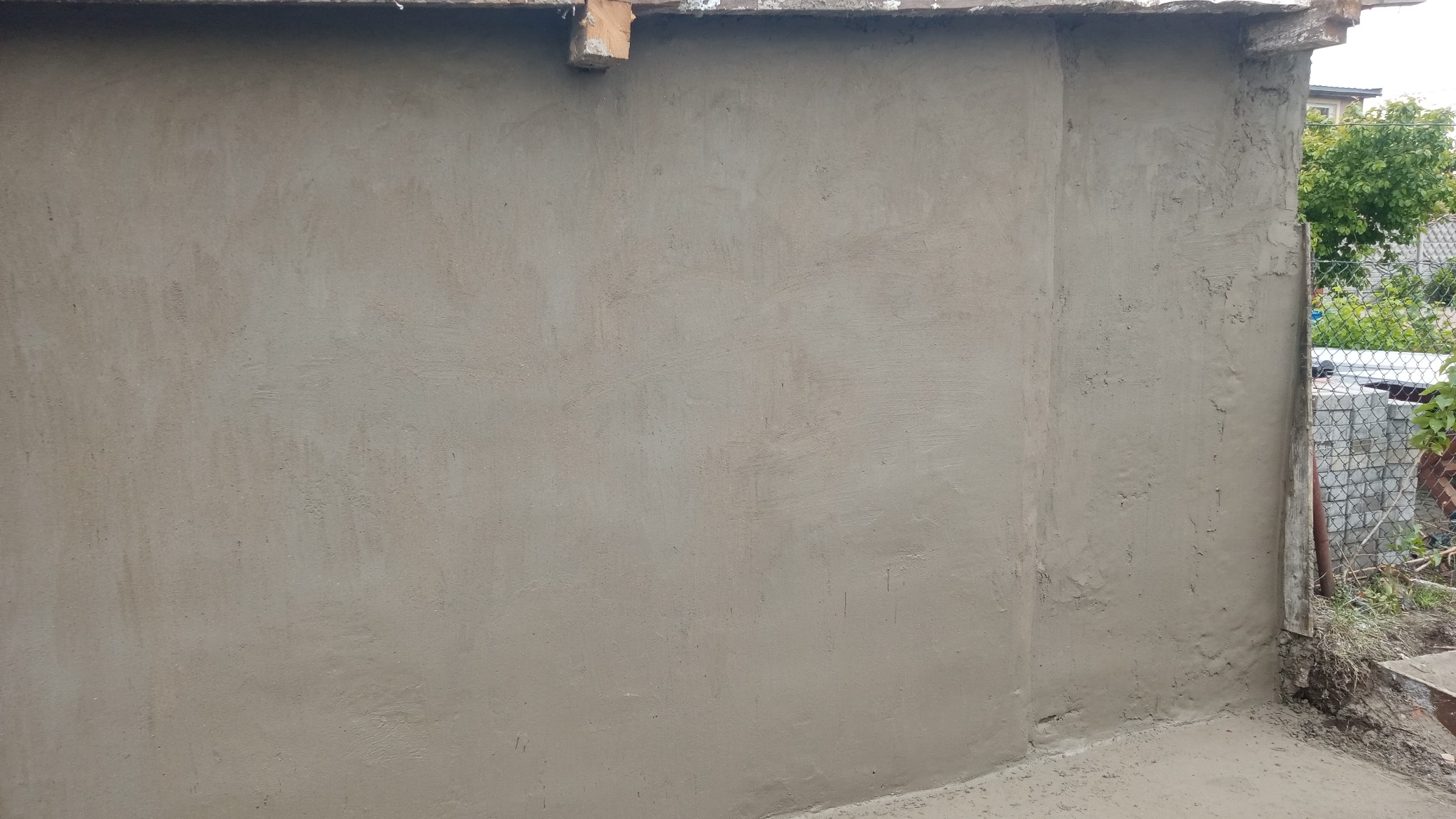 Świeżo otynkowana ściana zewnętrzna budynku z widocznym fragmentem dachu i otoczenia. Szara, gładka powierzchnia tynku.