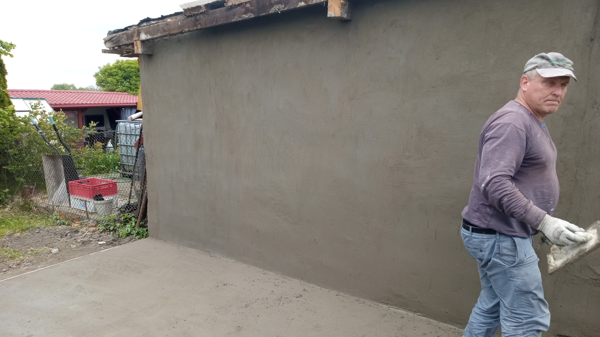 Świeżo otynkowana ściana budynku z widocznym fragmentem dachu i pracownikiem zacierającym tynk pacą. Ściana szara, tynk jeszcze wilgotny. W tle zieleń i zabudowania.