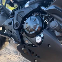 Czarny silnik motocykla Kawasaki z widocznymi detalami osprzętu, śrubami i logiem marki, sfotografowany z niskiego kąta na szutrowym podłożu.