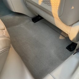 CarGlow Detailong - Czysta, szara wykładzina podłogowa w jasnym wnętrzu samochodu, widoczna spod fotela, z elementami siatki w tle. Oświetlenie słoneczne podkreśla detale.