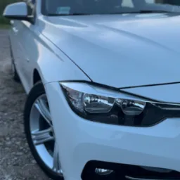 Białe BMW serii 3, widok z bliska na reflektor i koło. Auto stoi na żwirowej nawierzchni, w tle rozmyte drzewa i niebo o zachodzie słońca.
