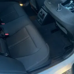 Czarne, tekstylne siedzenia tylne w samochodzie osobowym, widoczne zagłówki i pasy bezpieczeństwa. Biała karoseria na dole kadru.