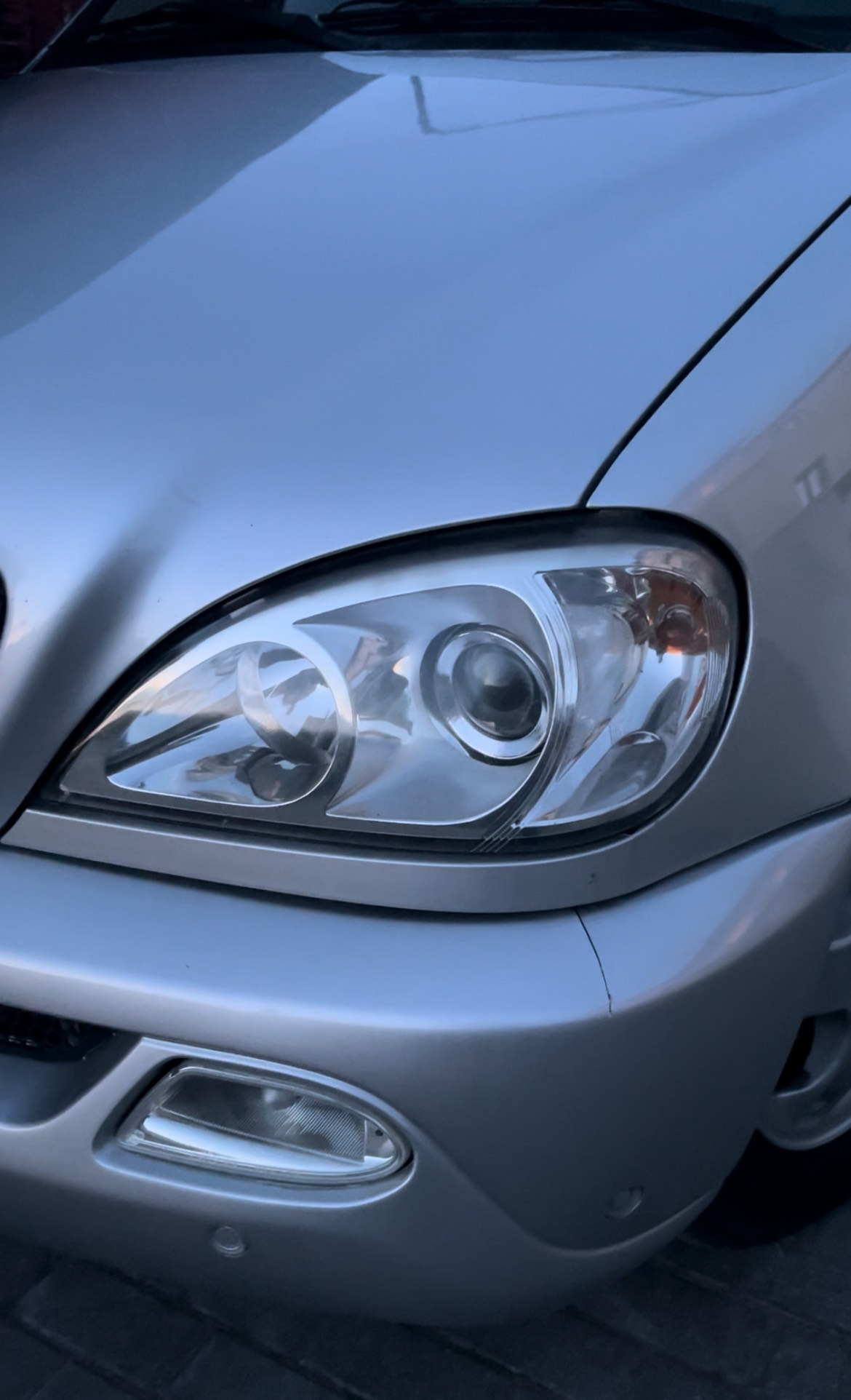 Srebrny samochód, zbliżenie na przedni reflektor i fragment maski. Widoczne detale lampy i zarys błotnika. Auto zaparkowane na zewnątrz.