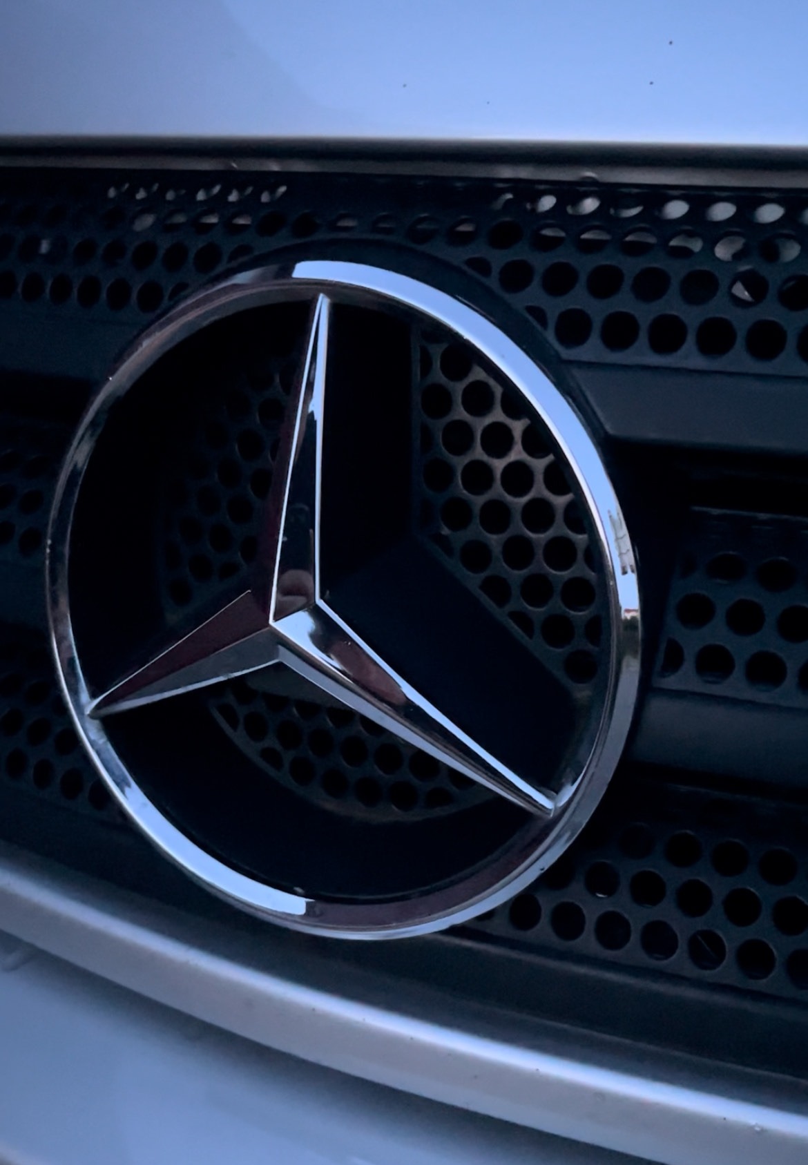 Błyszczące logo Mercedes-Benz na czarnej, perforowanej atrapie chłodnicy srebrnego samochodu, widziane z bliska pod kątem.