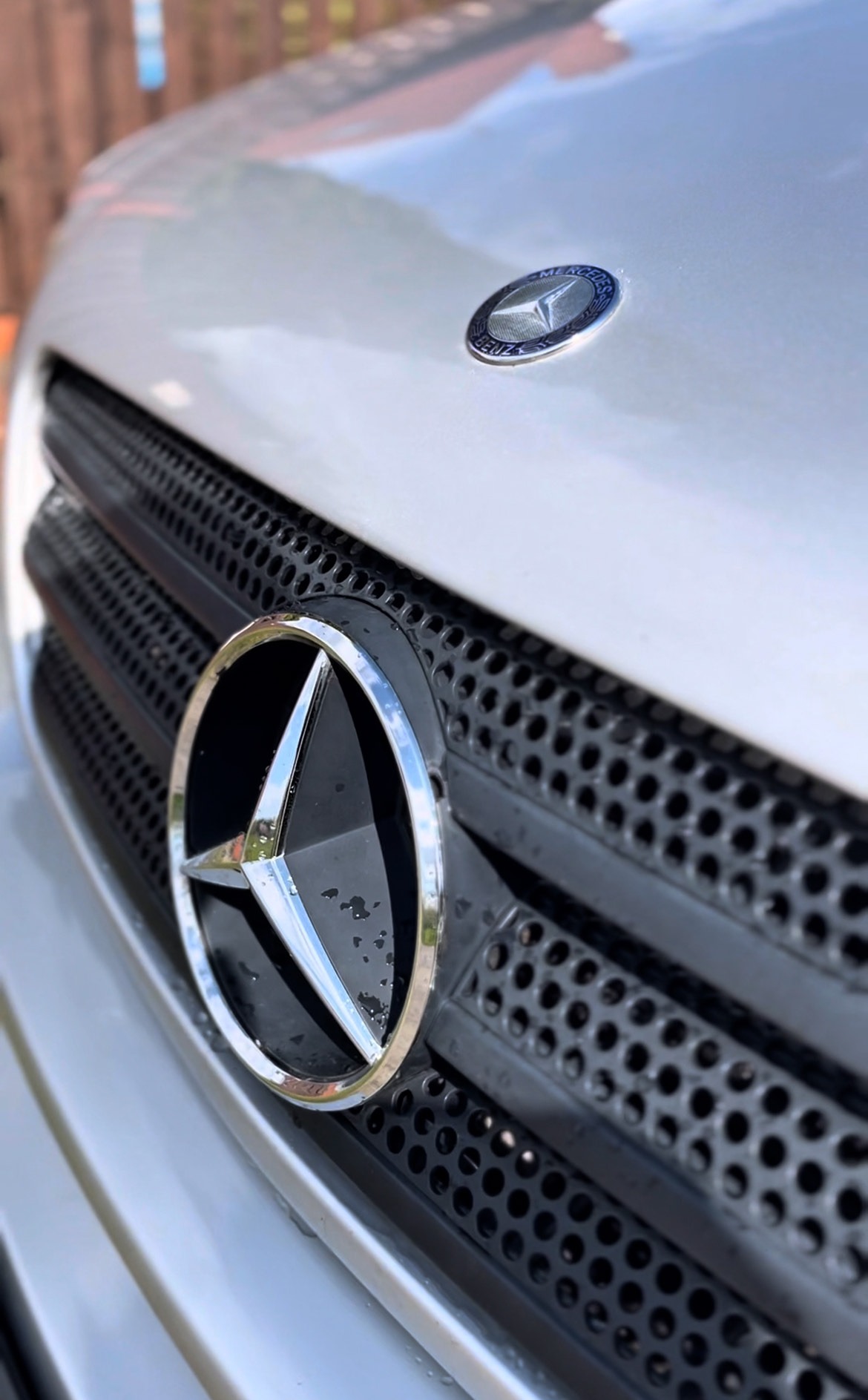 Błyszczący grill z logo Mercedesa, po umyciu. Widoczne krople wody. Chromowane obramowanie gwiazdy na tle czarnej siatki.