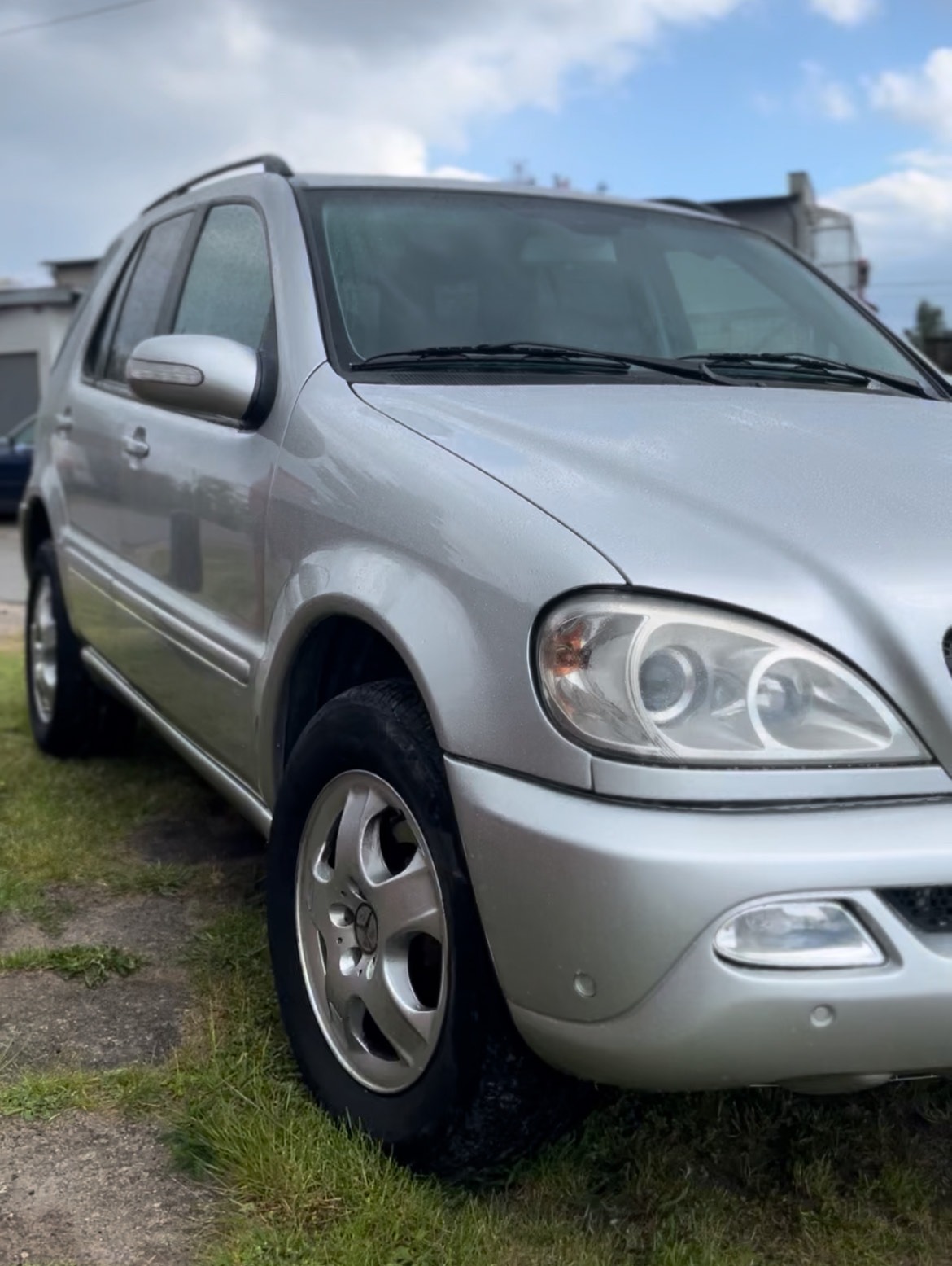 Srebrny SUV Mercedes-Benz ML W163 na trawie, widok z przodu, felgi aluminiowe z gwiazdą, lekko pochmurne niebo w tle. Auto po myjni.