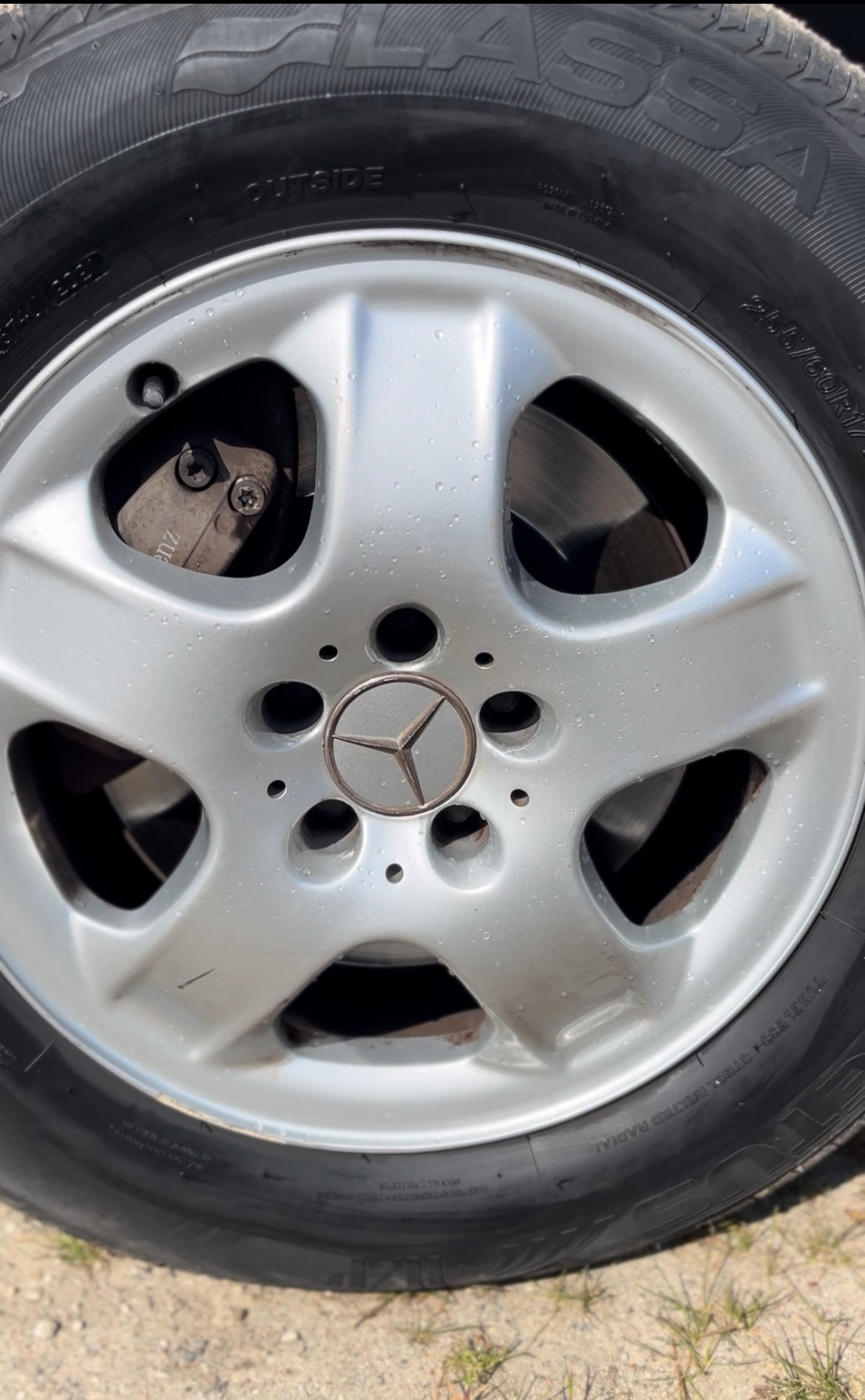 Błyszcząca, srebrna felga aluminiowa z logo Mercedesa, pokryta kroplami wody, zamontowana na oponie. Widoczny zacisk hamulcowy i fragment tarczy.