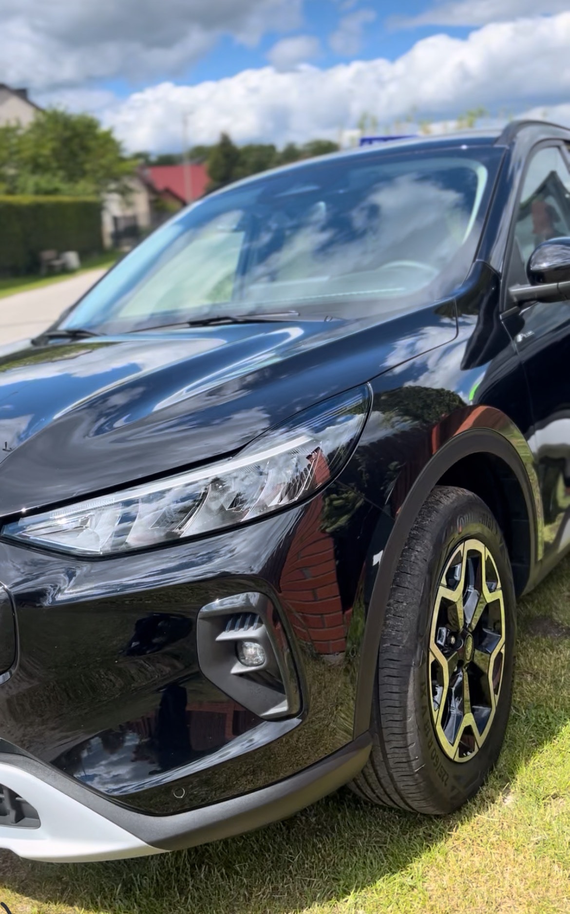 Lśniący, czarny SUV po myciu, z widocznym odbiciem nieba i chmur na karoserii. Detale felg i reflektorów podkreślone przez światło słoneczne. Auto stoi na trawie.