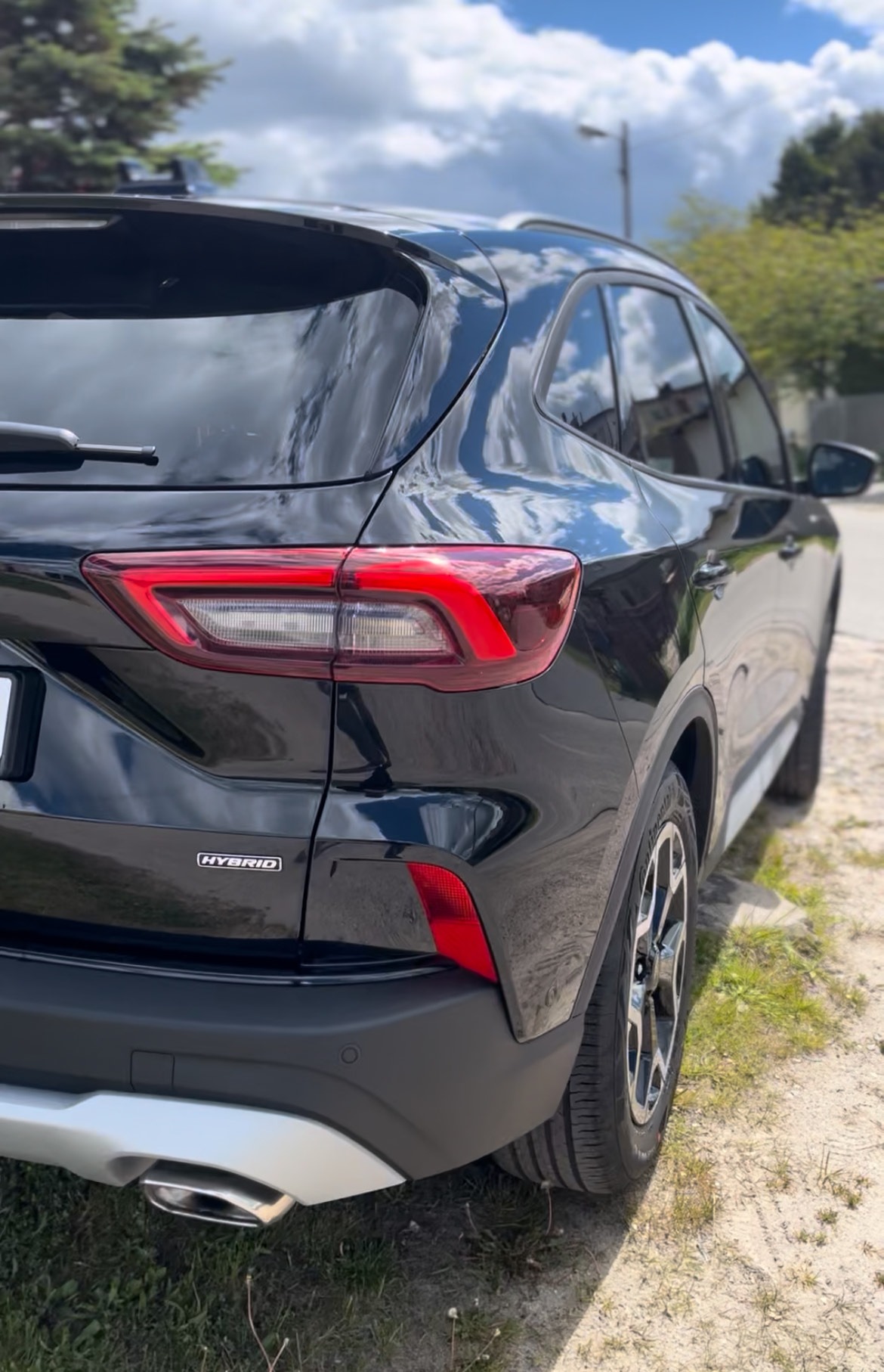 Błyszczący, czarny SUV Hybrid, widok z tyłu, z detalami lamp, felg i logo. Auto zaparkowane na poboczu, tło: zieleń i błękitne niebo.