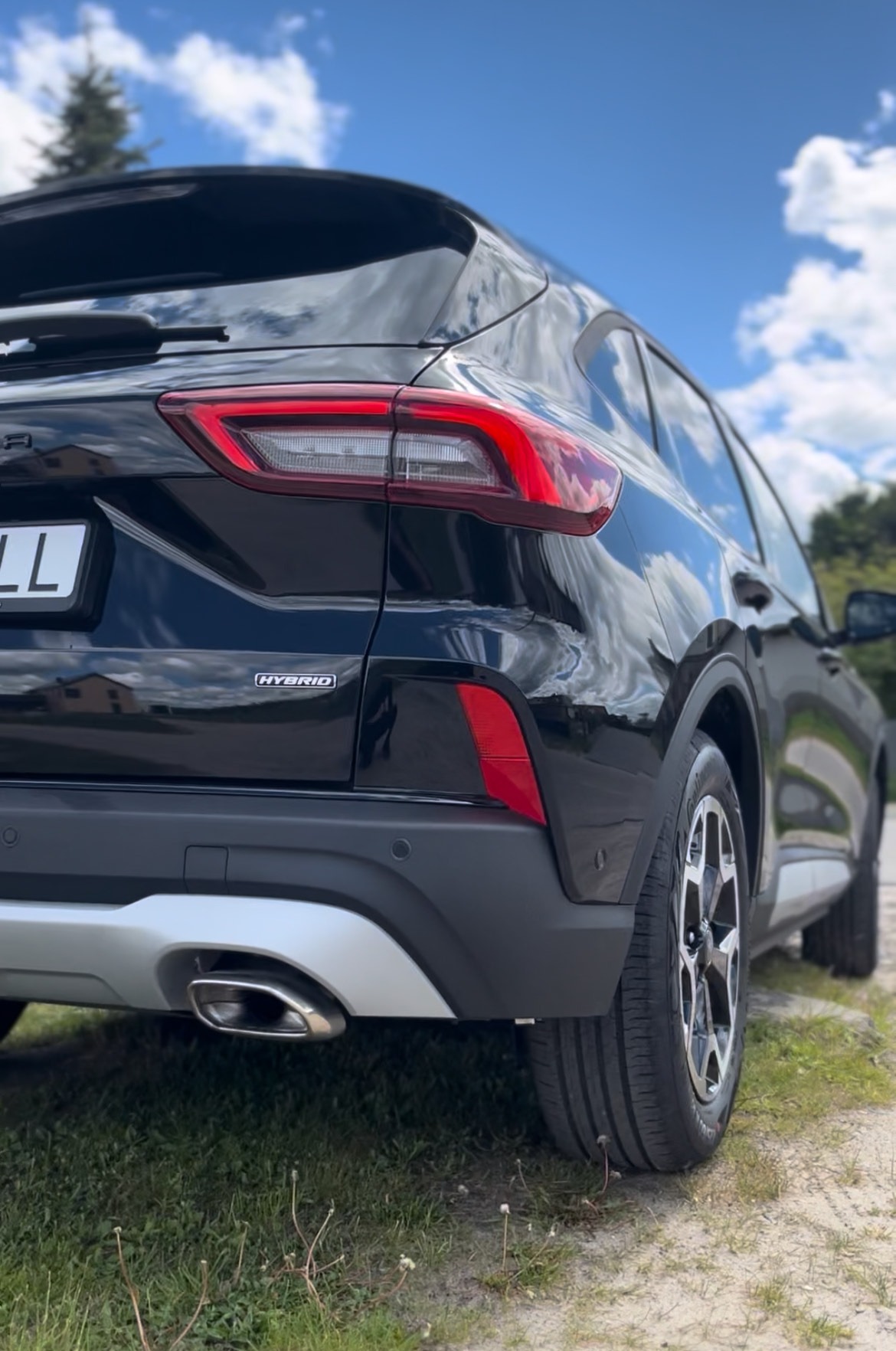 Błyszczący, czarny, hybrydowy SUV na trawie, widziany z tyłu pod kątem. Chromowany wydech i odbicie chmur w karoserii dodają elegancji.