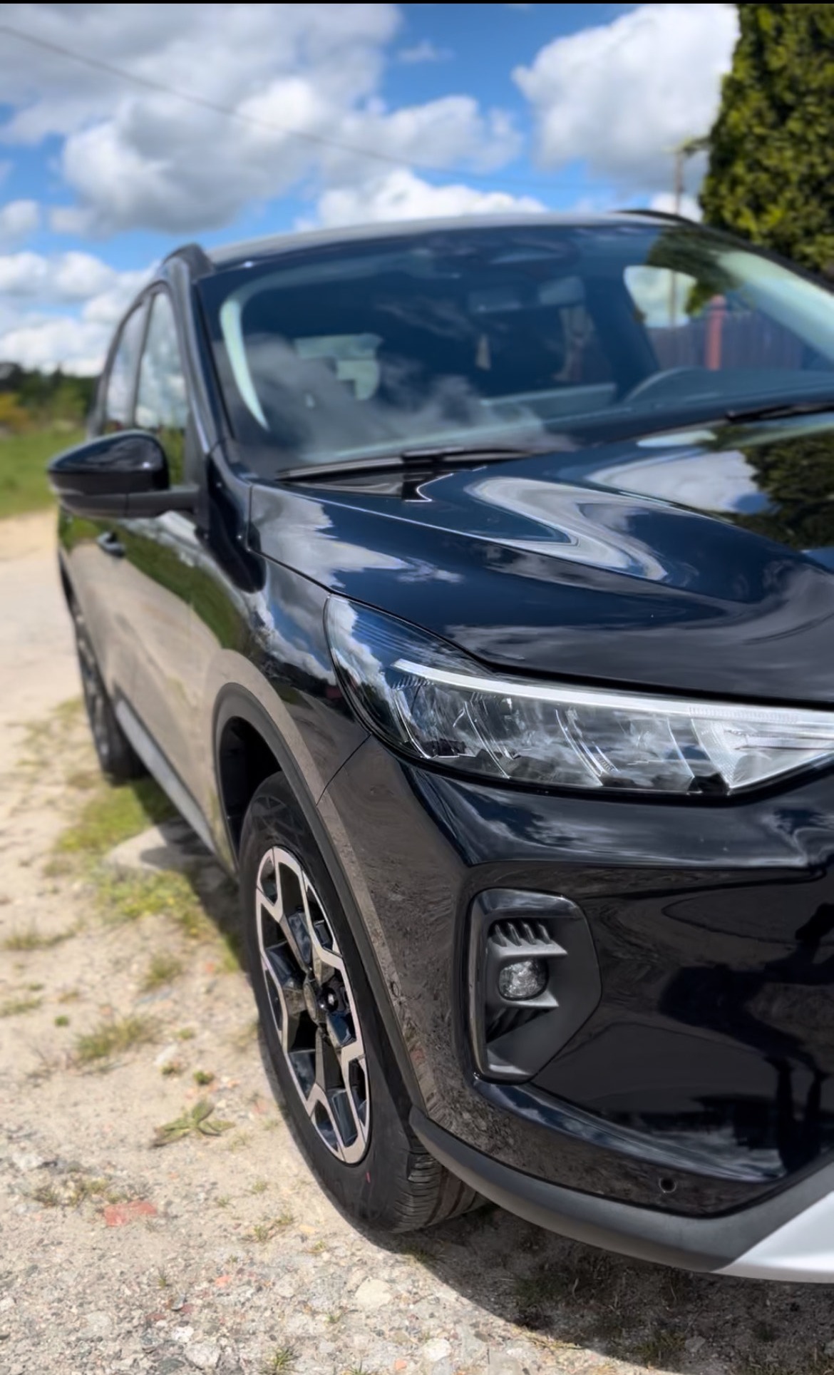 Błyszczący, czarny SUV na żwirowej drodze w słoneczny dzień. Widok z bliska na przedni reflektor i koło, z odbiciem chmur na karoserii.