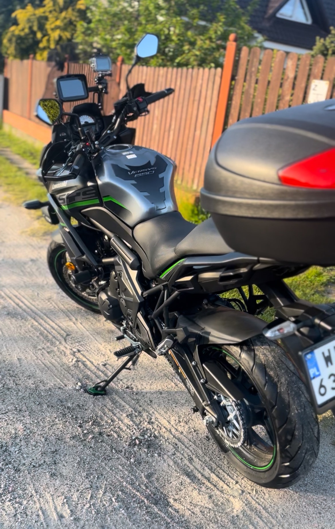 Motocykl Kawasaki Versys 650 z kufrem, zaparkowany na żwirowej drodze, z widocznym fragmentem drewnianego płotu w tle. Ujęcie z boku, w słoneczny dzień.