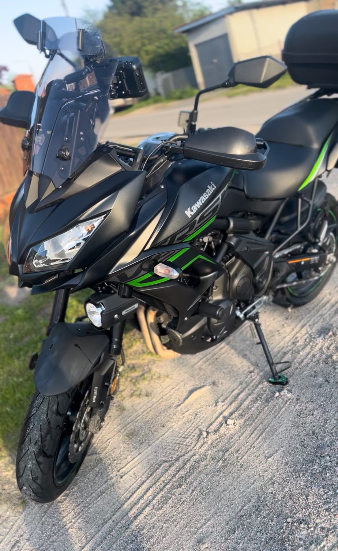 Czarny motocykl Kawasaki Versys 650 z zielonymi akcentami, zaparkowany na szutrowej drodze, z widocznymi elementami wyposażenia turystycznego.