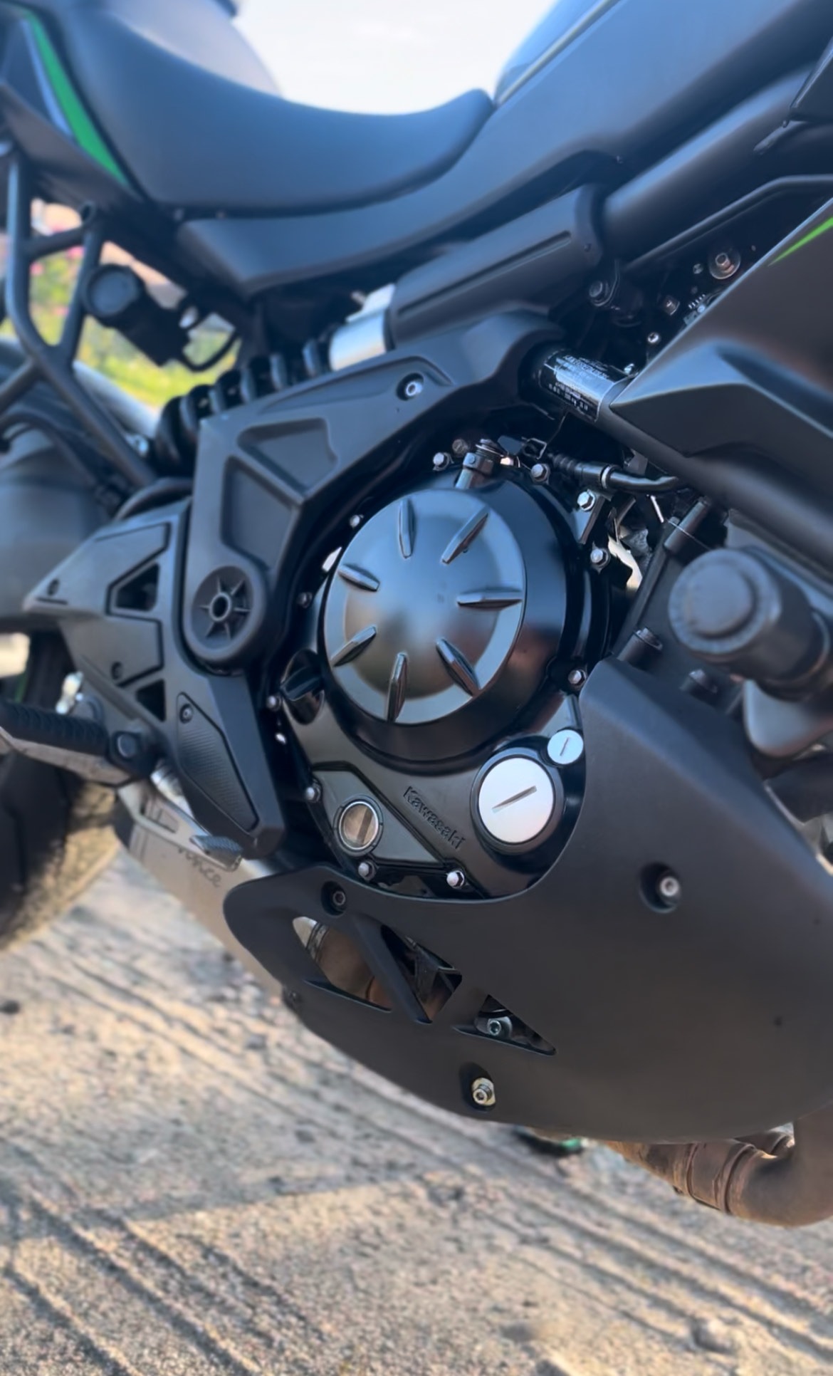 Czarny silnik motocykla Kawasaki z widocznymi detalami osprzętu, śrubami i logiem marki, sfotografowany z niskiego kąta na szutrowym podłożu.