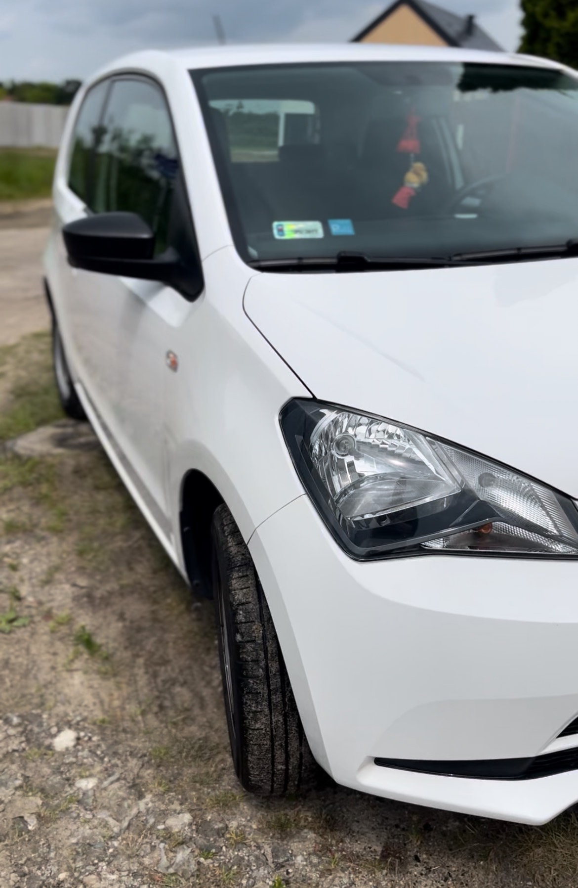 Biały samochód osobowy, widok z przodu na błotnik i reflektor, lekko zabrudzone opony, na tle zieleni i fragmentu domu.