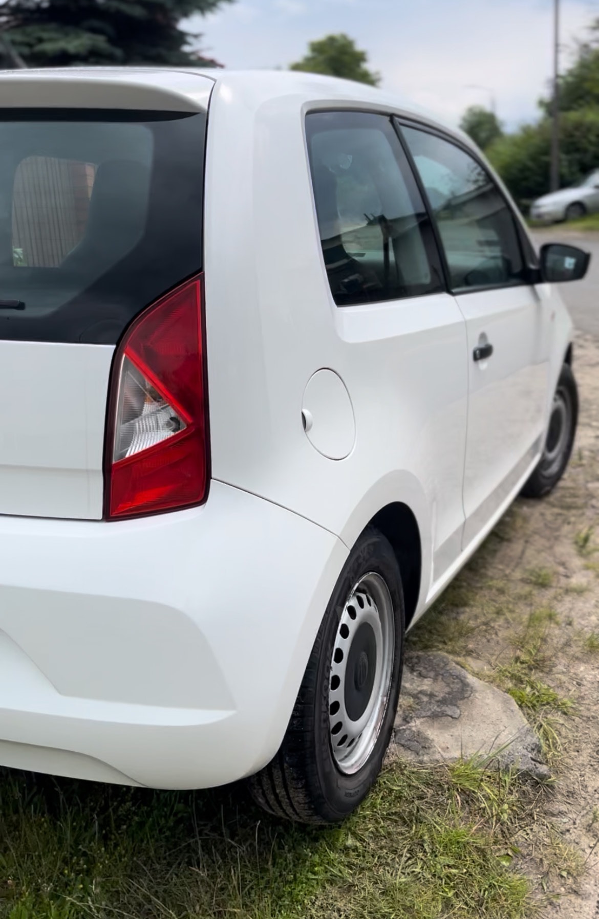 Biały samochód miejski z widocznym tyłem i kołem, stojący na poboczu z zieloną trawą. Widoczne detale karoserii i opon. Delikatne oświetlenie.