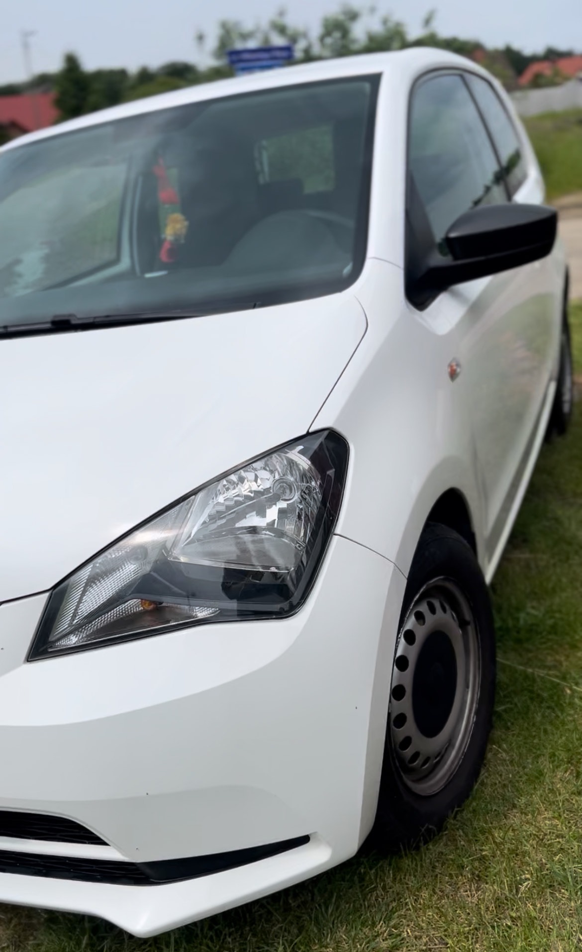 Biały samochód osobowy na trawie, widok z przodu na reflektor, koło i lusterko. W tle zieleń i błękitne niebo. Dekoracja w aucie.