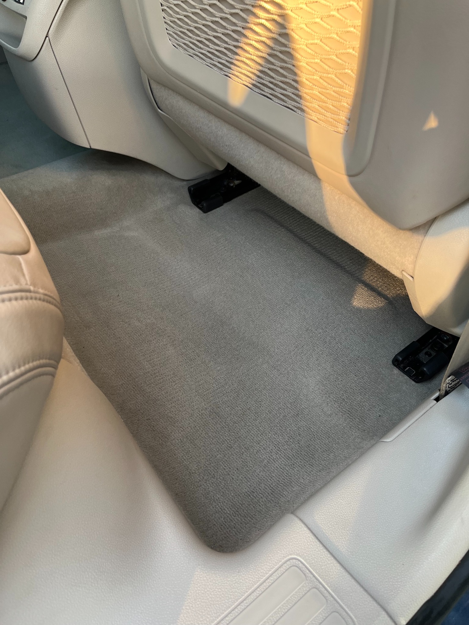 Czysta, szara wykładzina podłogowa w jasnym wnętrzu samochodu, widoczna spod fotela, z elementami siatki w tle. Oświetlenie słoneczne podkreśla detale.