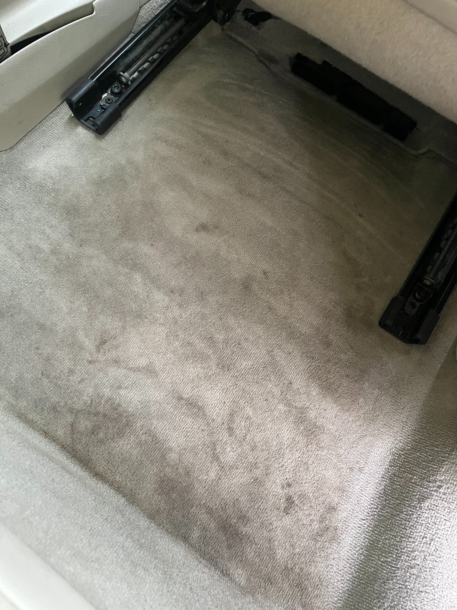 Szara, zabrudzona tapicerka pod fotelem samochodowym z widocznymi prowadnicami i śladami użytkowania. Delikatna faktura materiału i plamy podkreślają potrzebę czyszczenia.