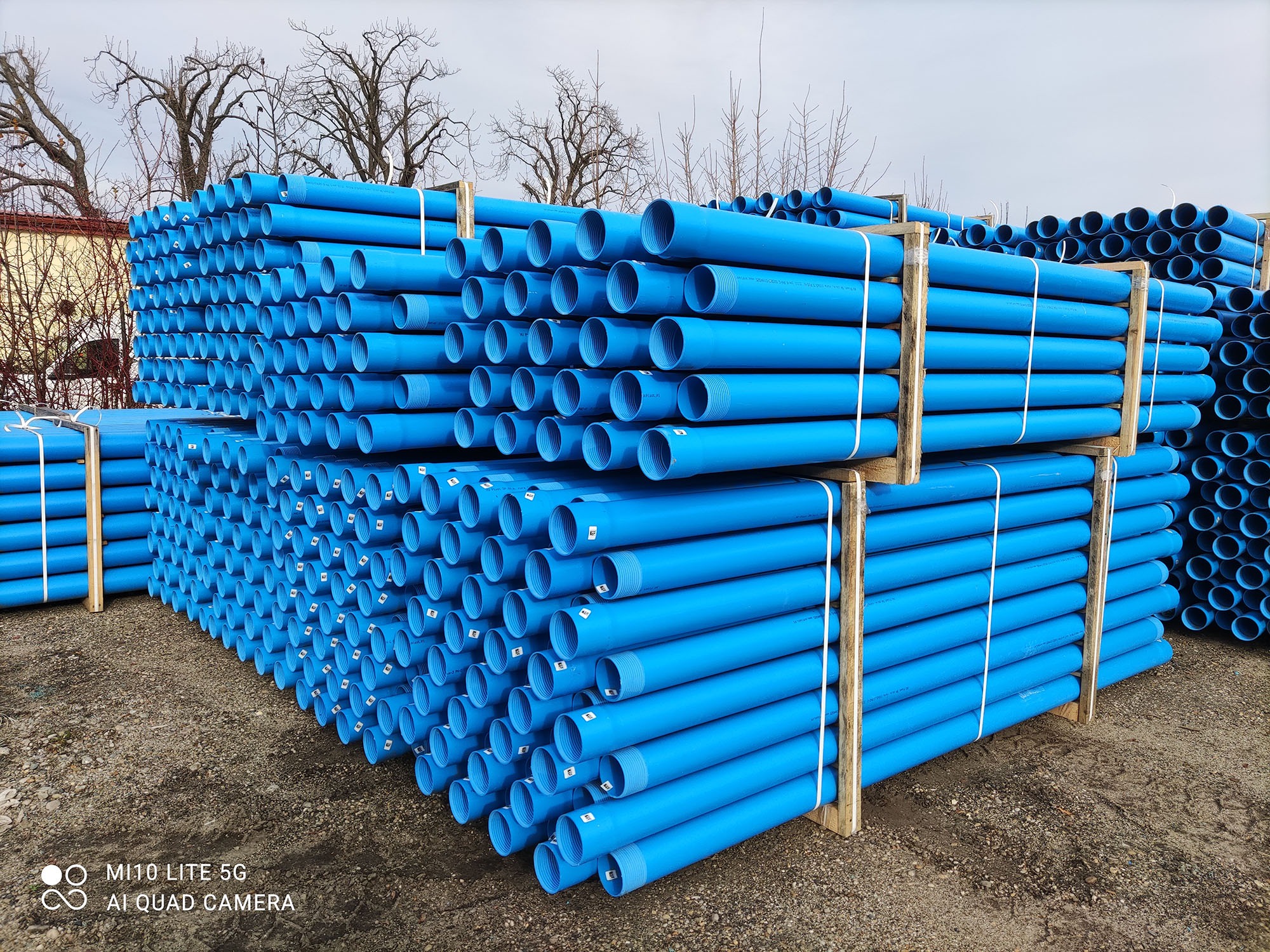 Stosy niebieskich rur PCV na paletach, widoczne gwintowane końcówki. Tło: budynek i drzewa bez liści. Rury do odwiertów studni głębinowych.
