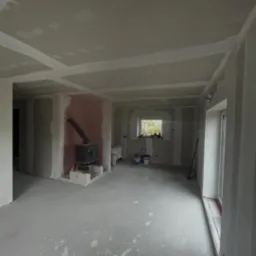 Wykończone ściany z płyt gipsowo-kartonowych w jasnym pomieszczeniu z piecem kaflowym. Widoczne łączenia płyt przygotowane do szpachlowania.