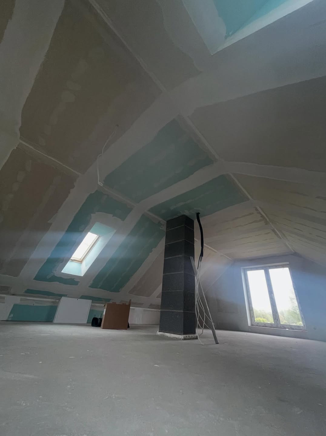 Poddasze w trakcie adaptacji: płyty gipsowe na skośnym suficie, widoczne łączenia i szpachlowanie, okno dachowe i komin w surowym wykończeniu.