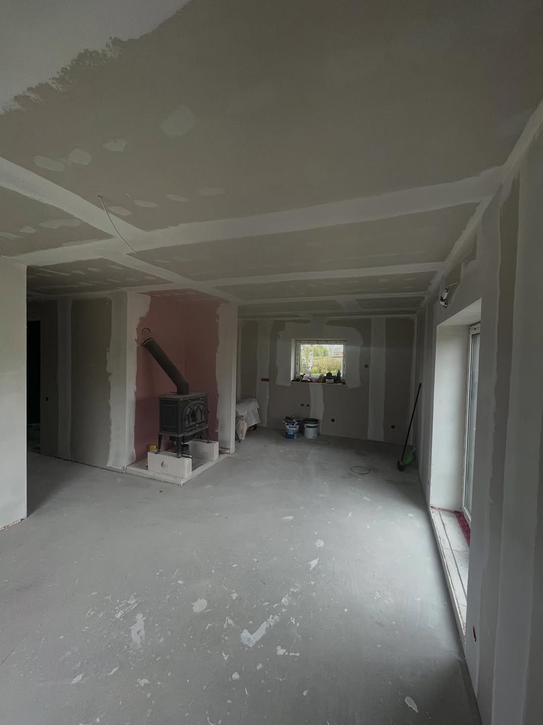 Wykończone ściany z płyt gipsowo-kartonowych w jasnym pomieszczeniu z piecem kaflowym. Widoczne łączenia płyt przygotowane do szpachlowania.