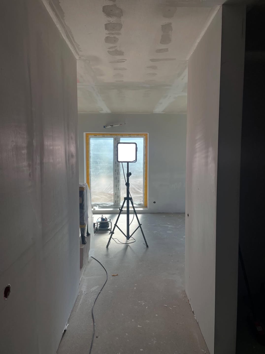 Wnętrze w trakcie szpachlowania: ściany przygotowane pod malowanie, widoczny reflektor na trójnogu, okno z żółtą ramą. Prace wykończeniowe w surowym stanie.