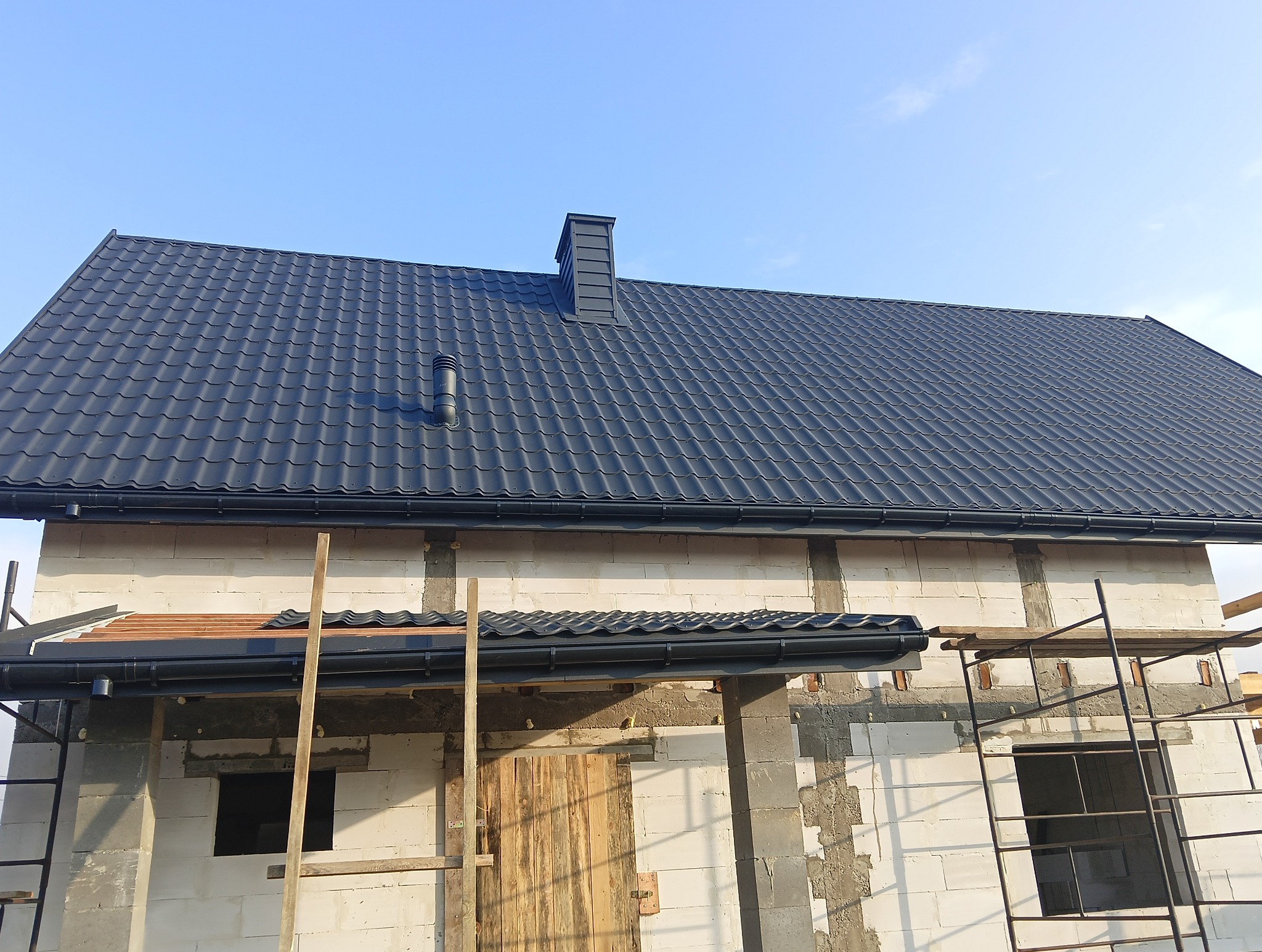 Nowy, grafitowy dach z blachodachówki z kominem i rynnami na surowym budynku z białego bloczka, w trakcie budowy, z widocznymi rusztowaniami.