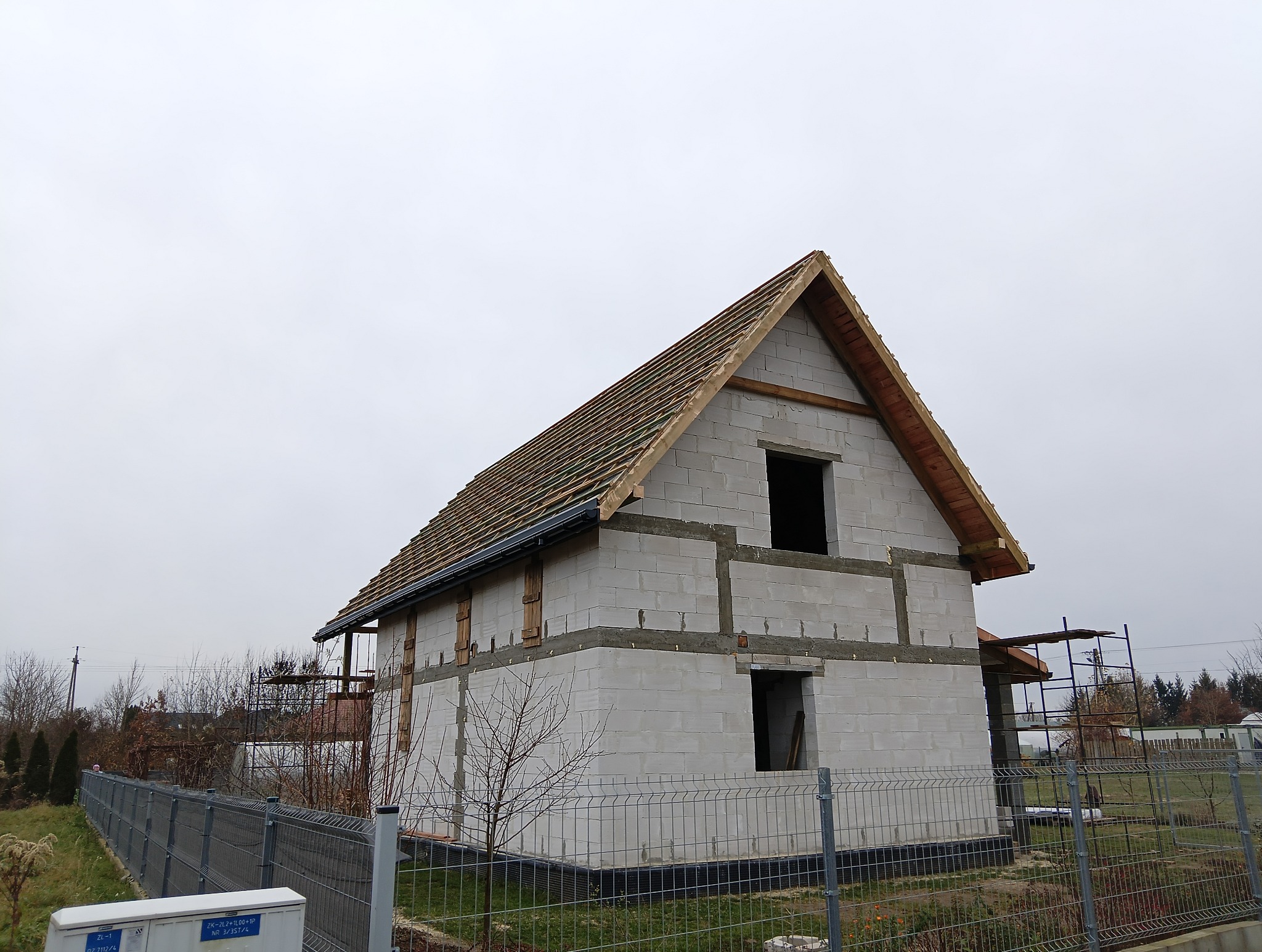 Budowa domu jednorodzinnego w stanie surowym otwartym. Widoczne ściany z bloczków, więźba dachowa i przygotowane otwory okienne. Szara, pochmurna pogoda.
