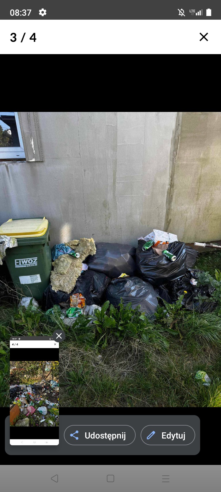 Sterta odpadów budowlanych i komunalnych w czarnych workach, obok zielonego pojemnika na śmieci, przy szarej ścianie budynku. Widoczne fragmenty zieleni.