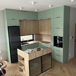 skleppodlogi.pl - Nowoczesna kuchnia w odcieniach zieleni i drewna z wyspą, czarnym sprzętem AGD i parkietem ułożonym w jodełkę. Widok na całe pomieszczenie, oświetlenie punktowe.