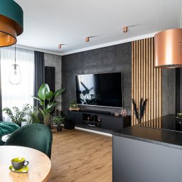 skleppodlogi.pl - Nowoczesny salon z drewnianą podłogą, zielonymi akcentami i ścianą z dekoracyjnymi panelami. Centralnie umieszczony telewizor i designerski okap.