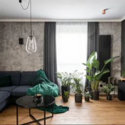 Stylowy salon z drewnianą podłogą, szarą sofą narożną, czarnym stolikiem i licznymi roślinami doniczkowymi. Ściana z efektem betonu i czarny grzejnik.